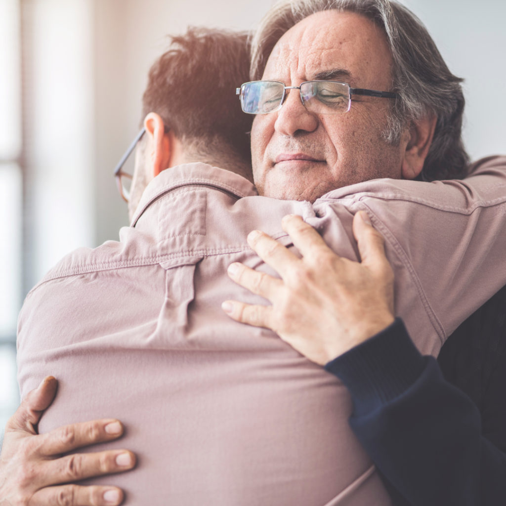 Middle aged man hugs his adult son. He looks supported.